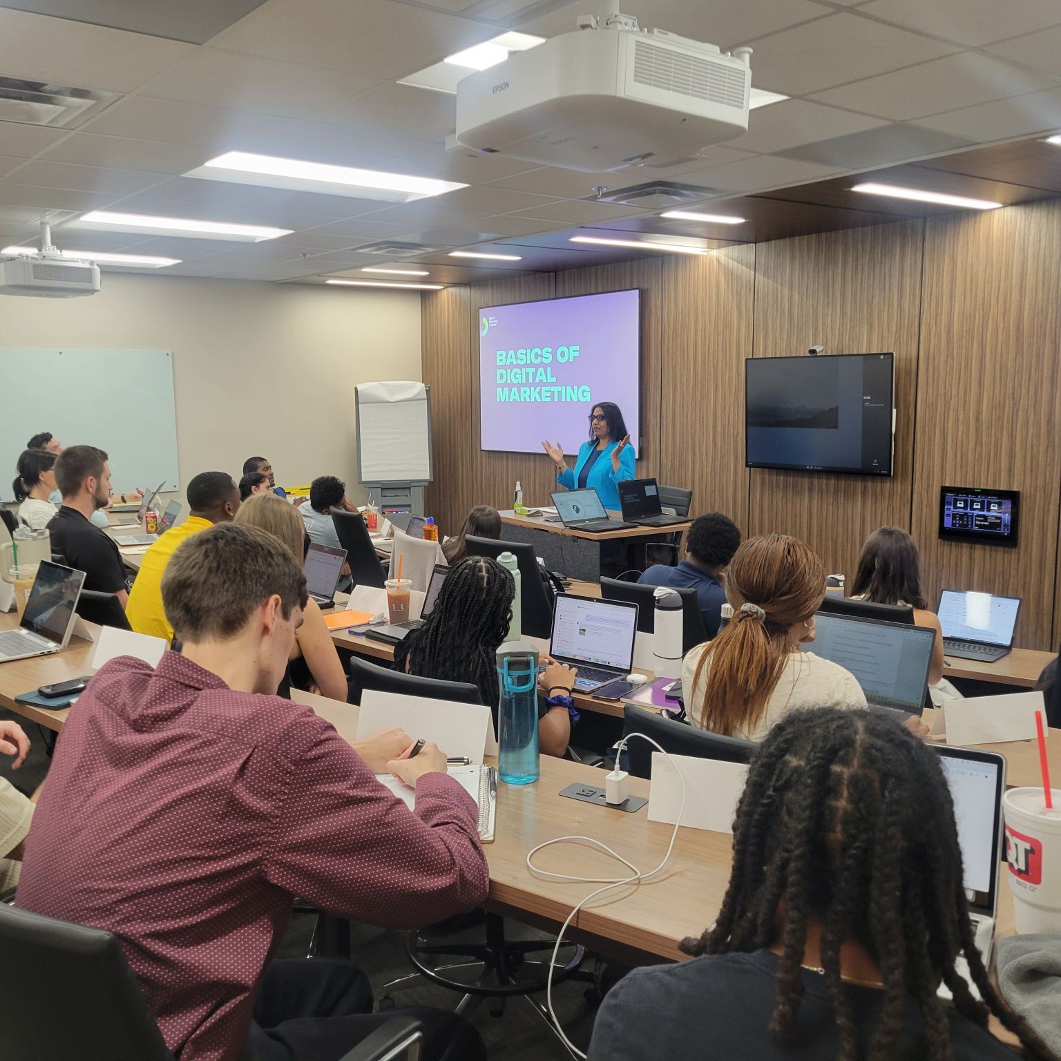 A female instructor presents "Basics of Digital Marketing" to a classroom of students with laptops.