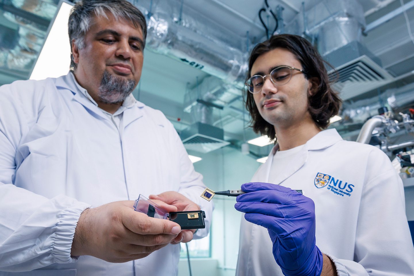 Two scientists in lab coats examine microchips. One holds a chip with tweezers, the other holds a container.
