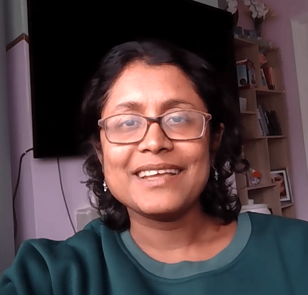 Smiling woman in glasses and green top, dark hair, with TV and bookshelf in background.