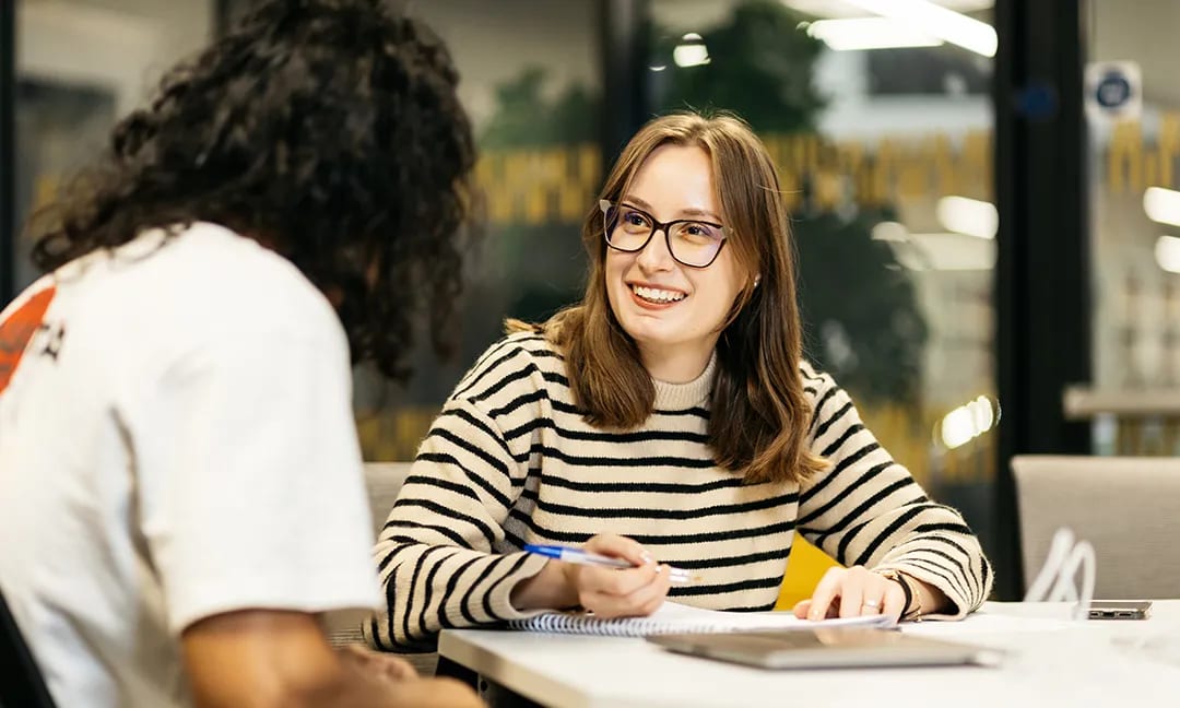 Smiling woman in glasses and striped sweater writes, talking to another person at a table.