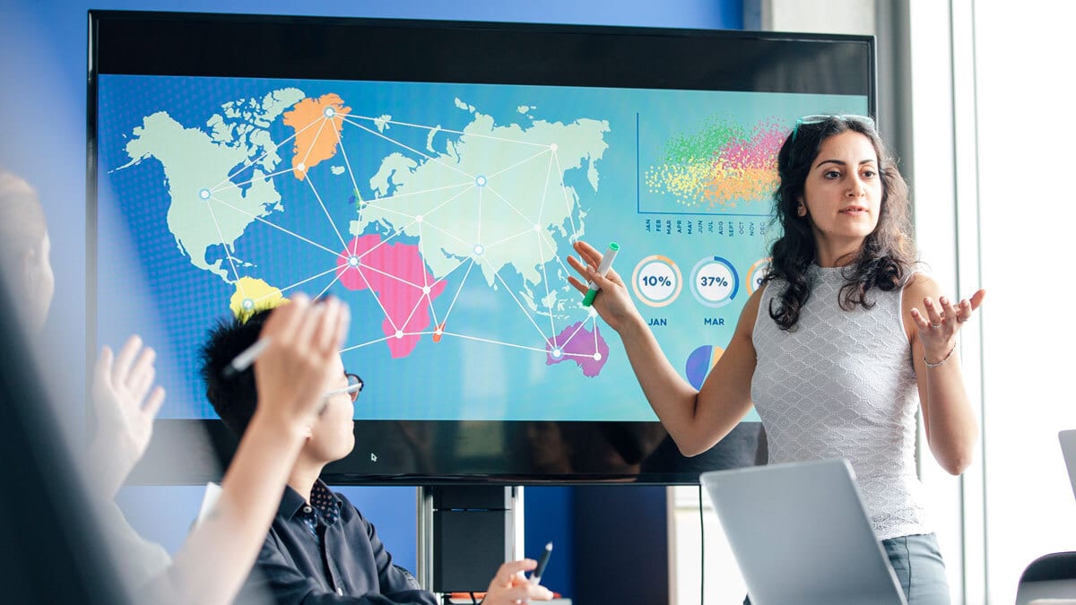 Woman presenting data visualizations and a world map on a large screen to colleagues.