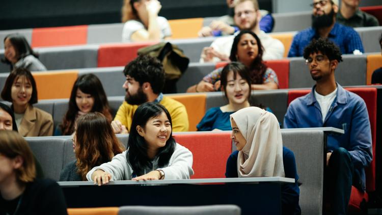 Diverse university students in a tiered lecture hall, some smiling and interacting.
