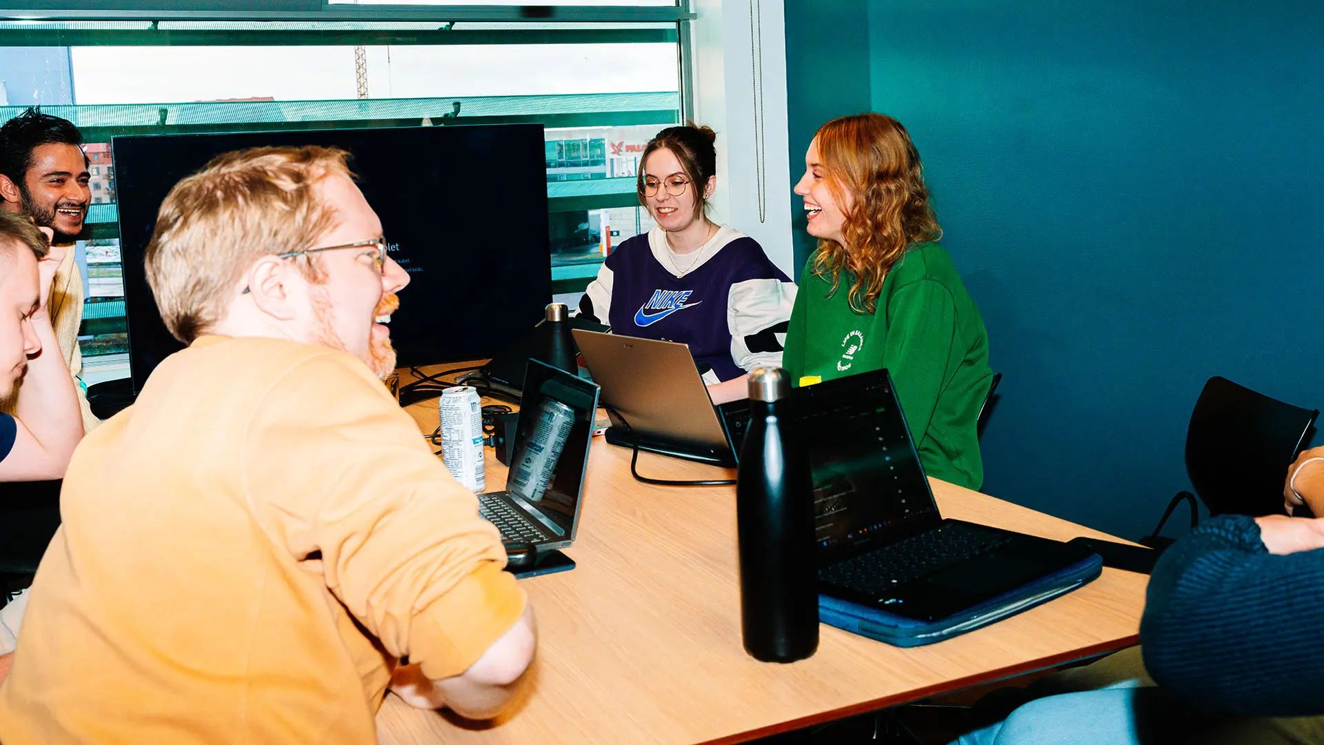 Smiling young people collaborate around laptops in a bright, modern meeting room.
