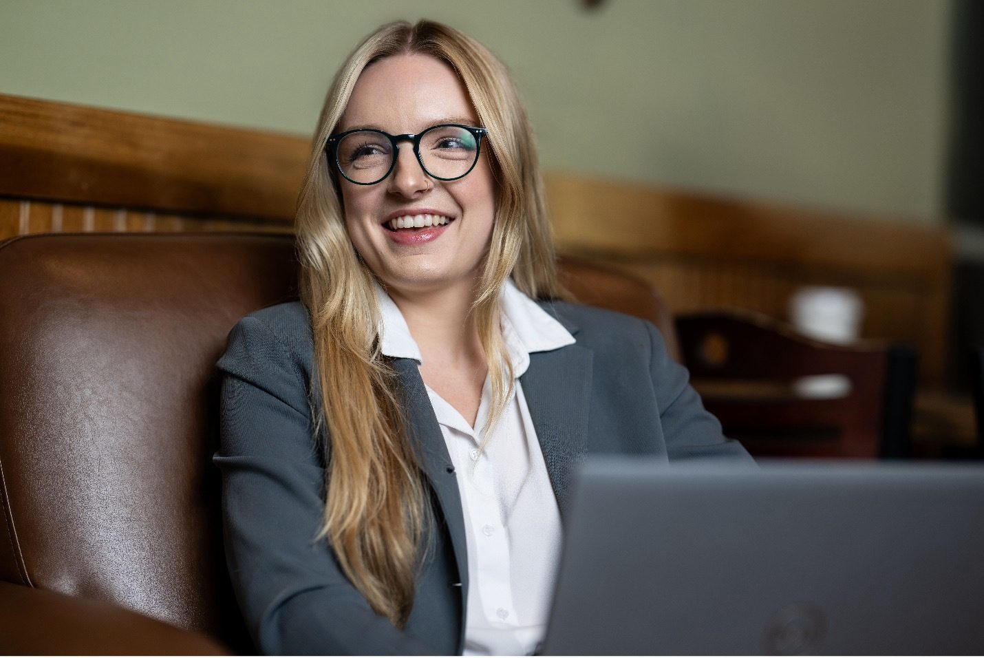 A smiling blonde woman in glasses and a suit jacket, seated, looking right, with a laptop nearby.