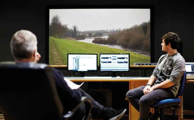 Two men in a studio watch a large screen with a river landscape and computer monitors.