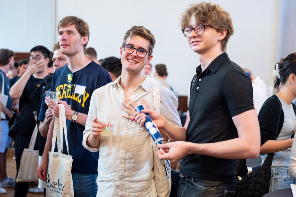 Three young men smiling at a social event, holding drinks and a blue tube.