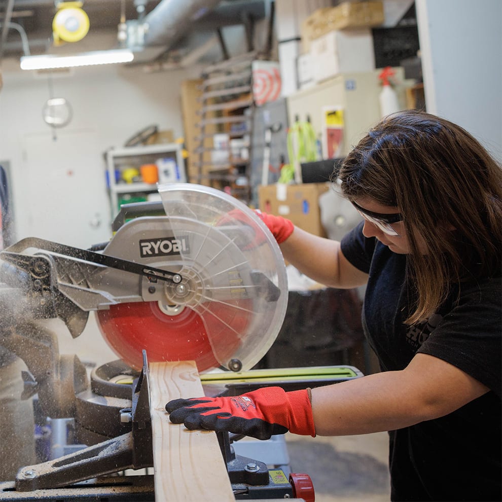 A woman in safety glasses and gloves uses a miter saw to cut a piece of wood in a workshop.