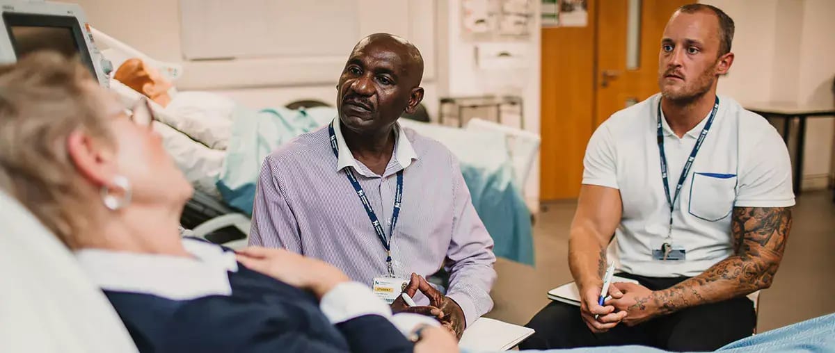Two male students observe a female patient in a medical training room.
