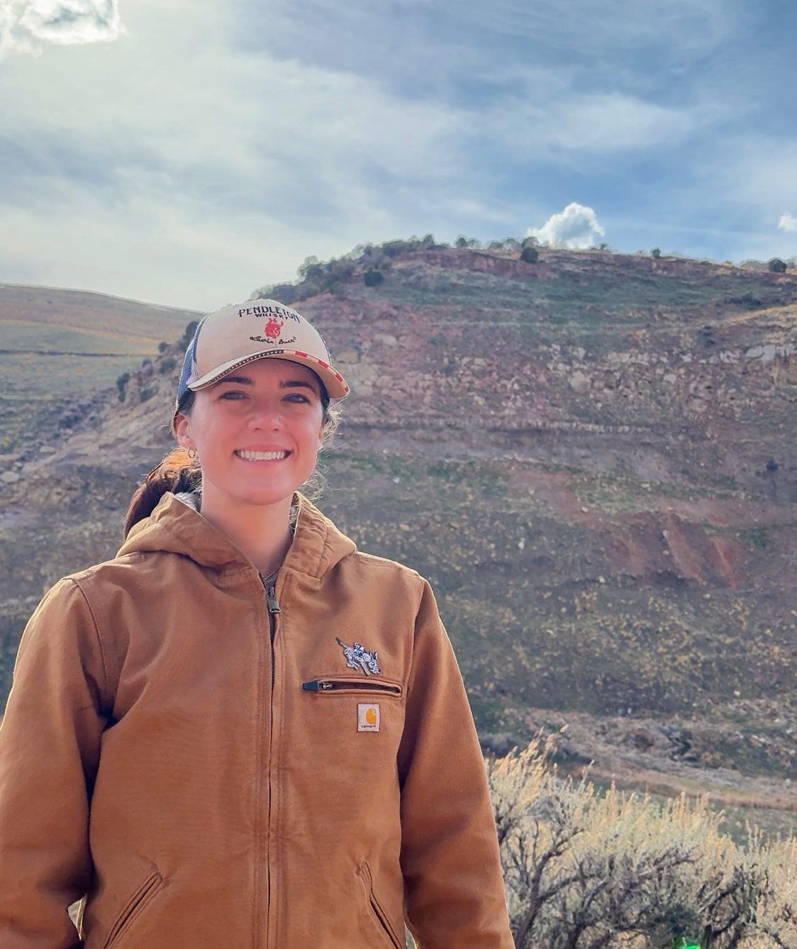 Smiling woman in a Pendleton cap and brown Carhartt jacket, mountains in the background.