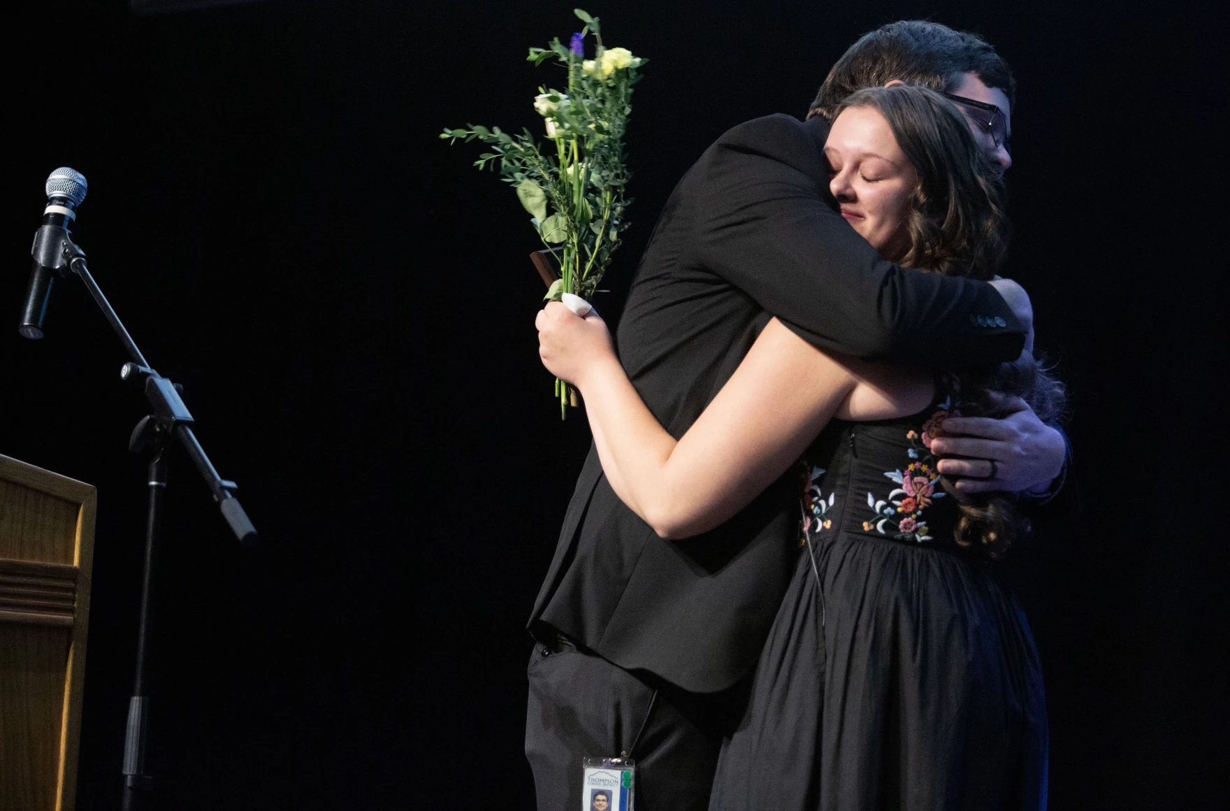 A man and a woman embrace on a dark stage; she holds a bouquet of flowers and wears an embroidered black dress.