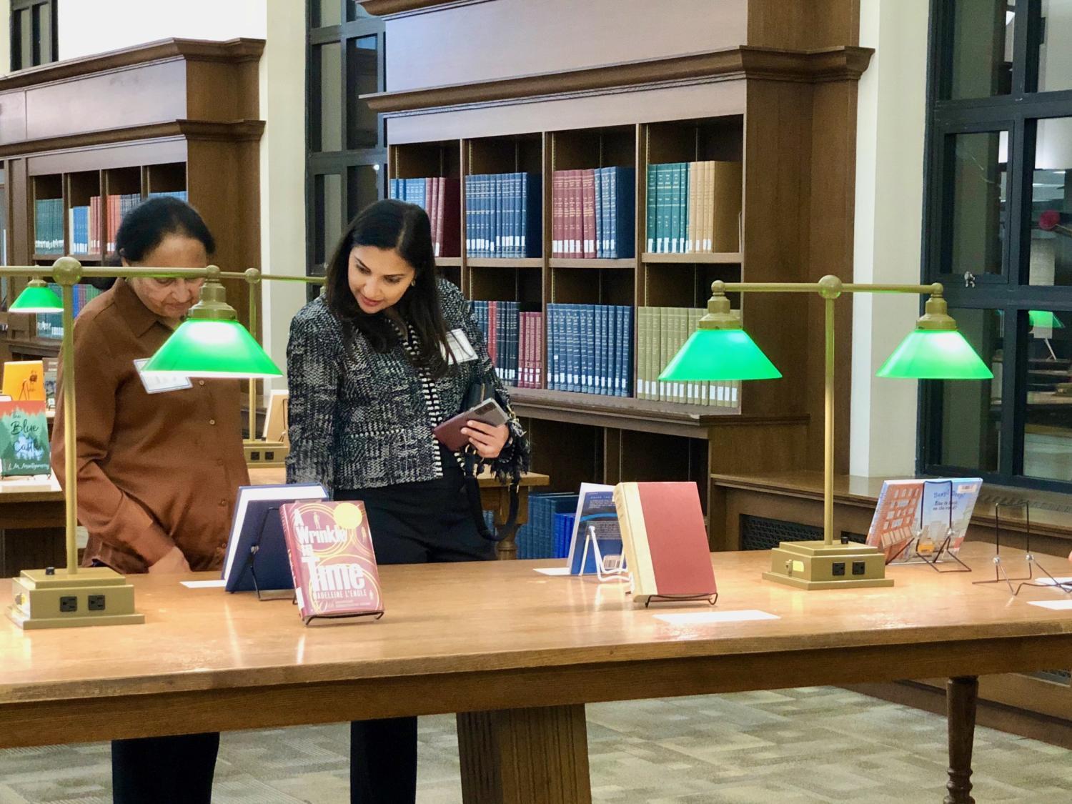 Two women browse books in a library with green desk lamps.