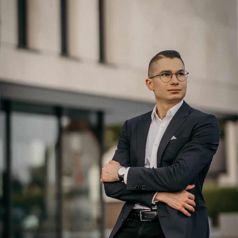 Man in a suit and glasses, arms crossed, looking to the right.