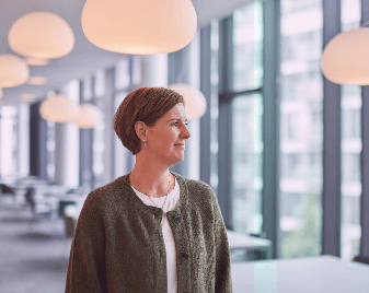 A woman in a green cardigan stands in a modern office with large windows, looking thoughtfully right.