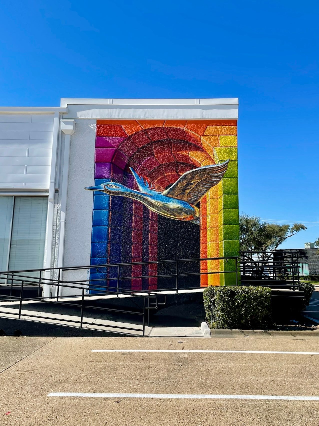 Vibrant mural of a blue bird soaring over a rainbow-colored sun on a white building, bright blue sky.