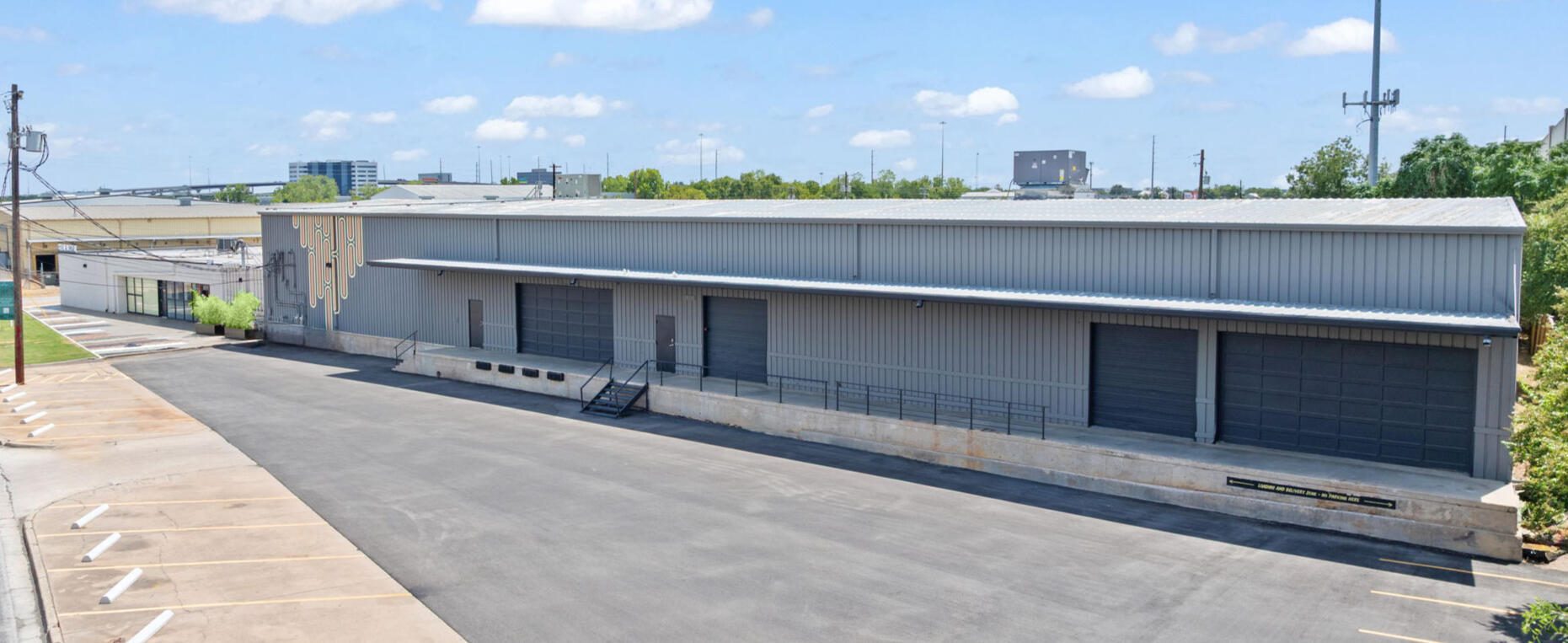 A long, grey industrial building with multiple loading docks, a large paved area, and a decorative wall pattern.