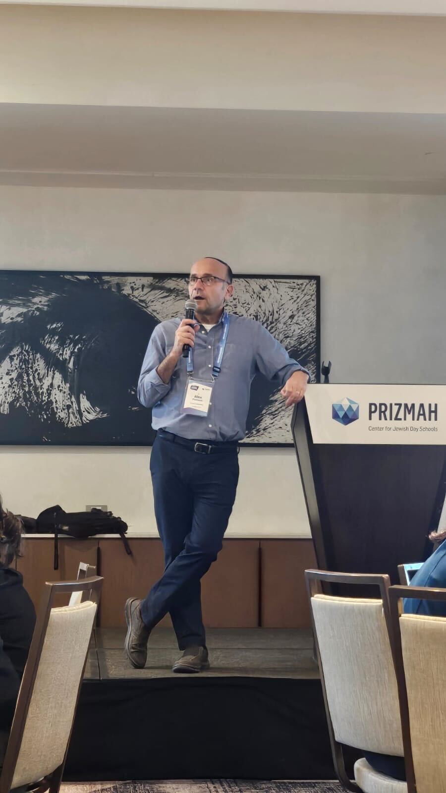 Man with glasses speaking into a microphone on a stage, leaning on a PRIZMAH podium.