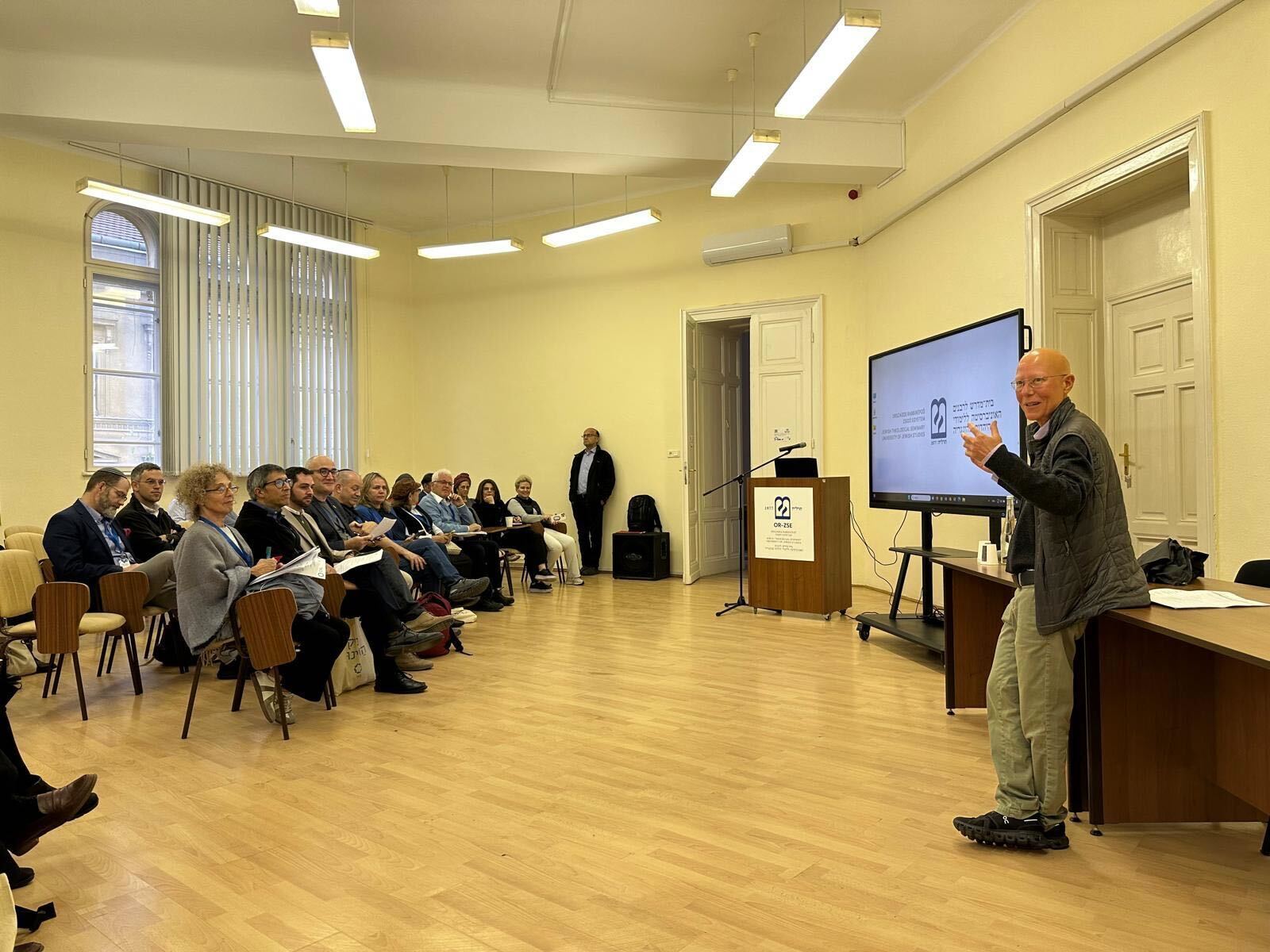 A speaker gestures to an audience in a lecture hall, next to a large screen displaying a logo.