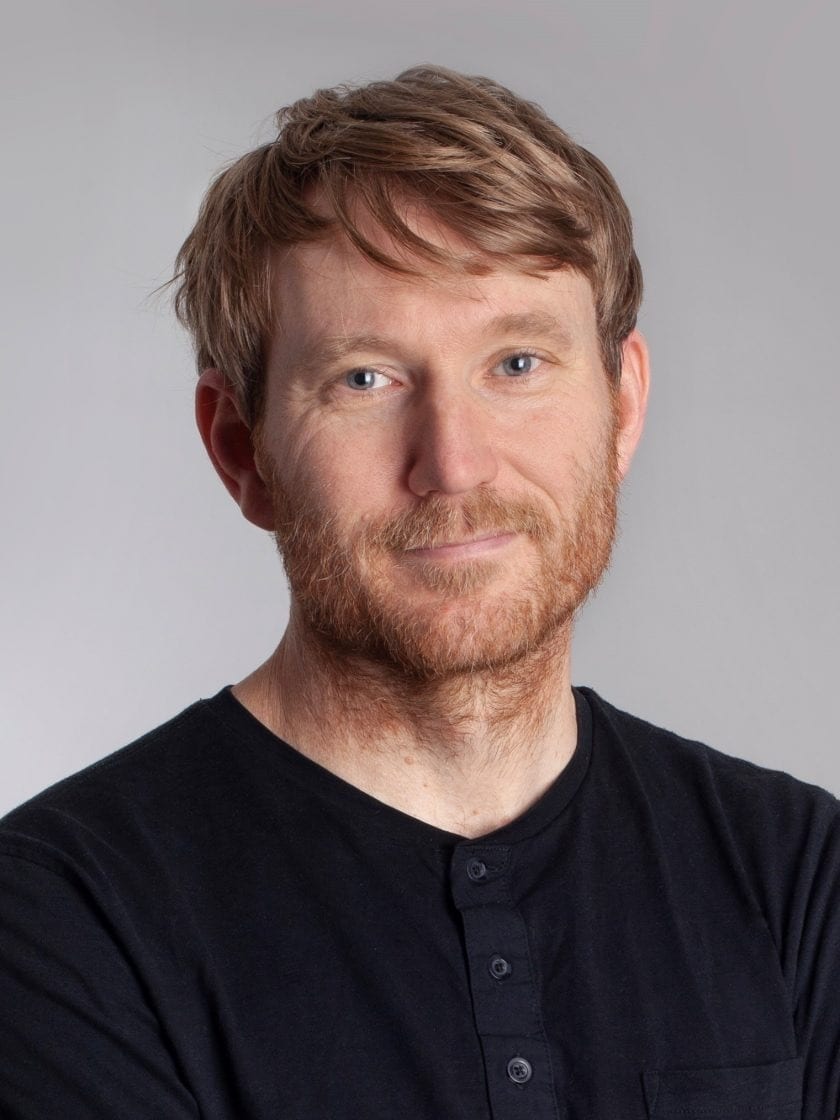 Smiling man with red hair, beard, blue eyes, black henley shirt on gray background.