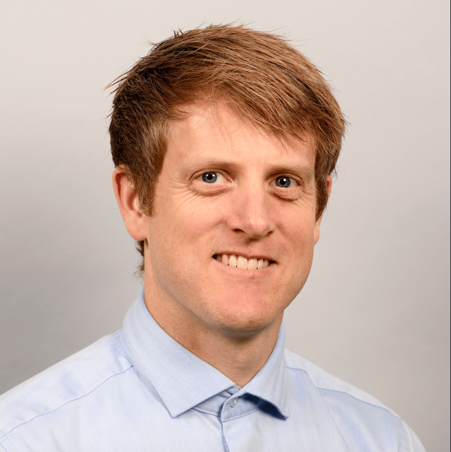 Headshot of a smiling man with reddish-brown hair and blue eyes, wearing a light blue shirt.