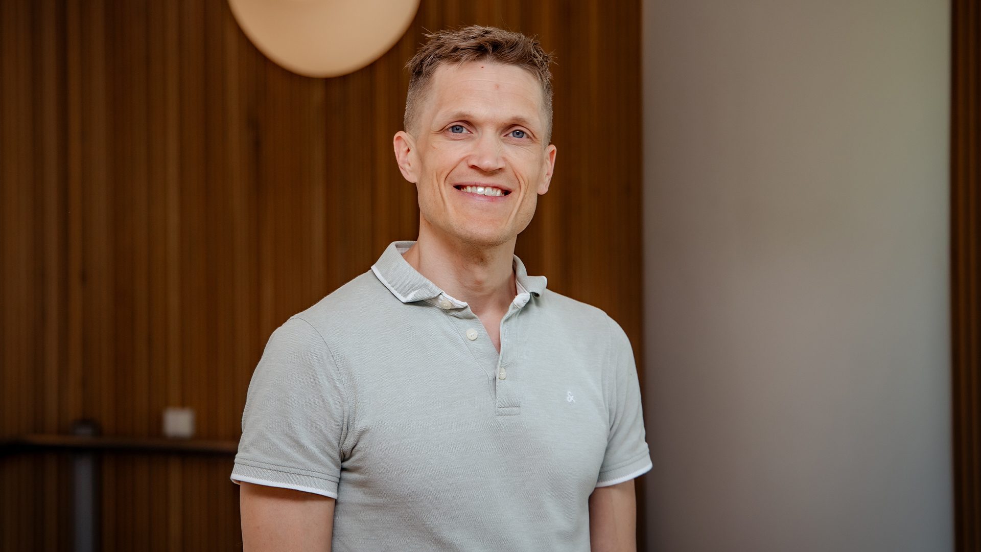 Smiling man in a light green polo shirt looking at the camera against a wood and light wall background.