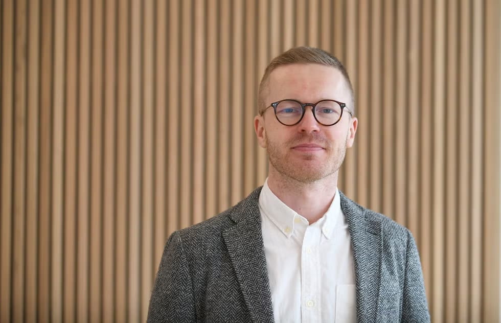 A man with glasses and a tweed jacket smiles faintly in front of a wooden slatted wall.
