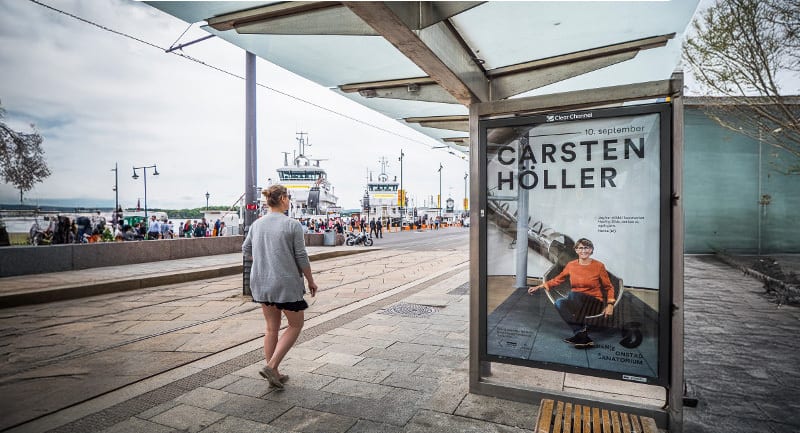 A woman walks past a bus stop with a "Carsten Höller" poster, overlooking a bustling harbor with ships.