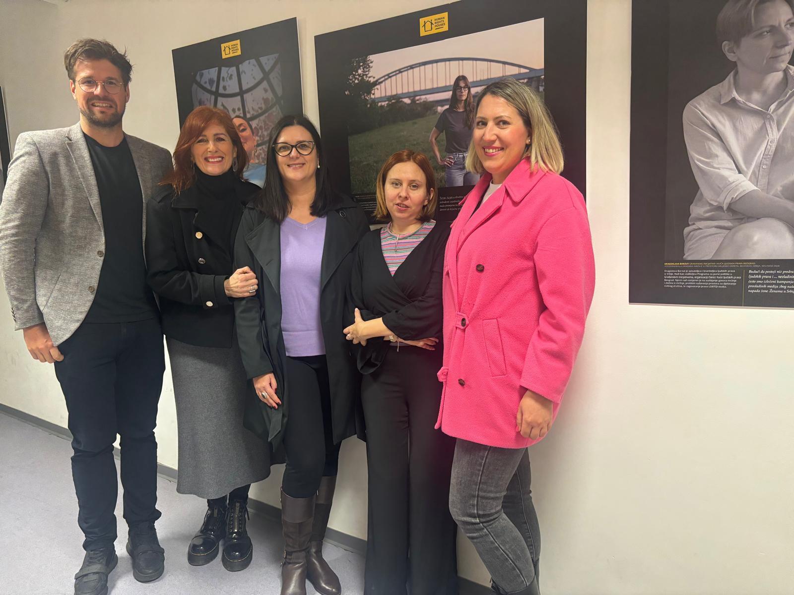 Five people, three women and two men, standing in a line in front of artwork.