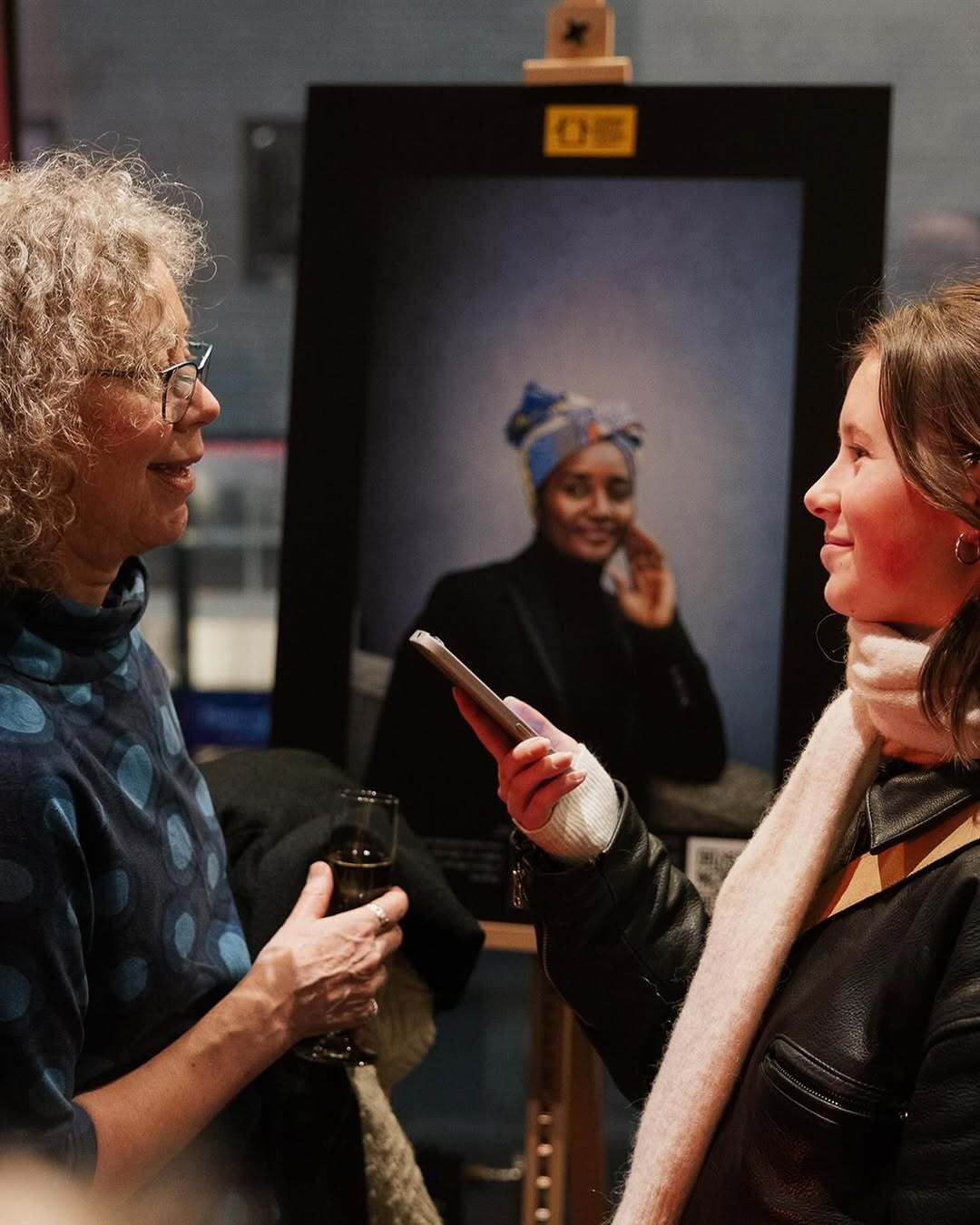 Two women conversing, one holding champagne, the other a phone, with a portrait in the background.