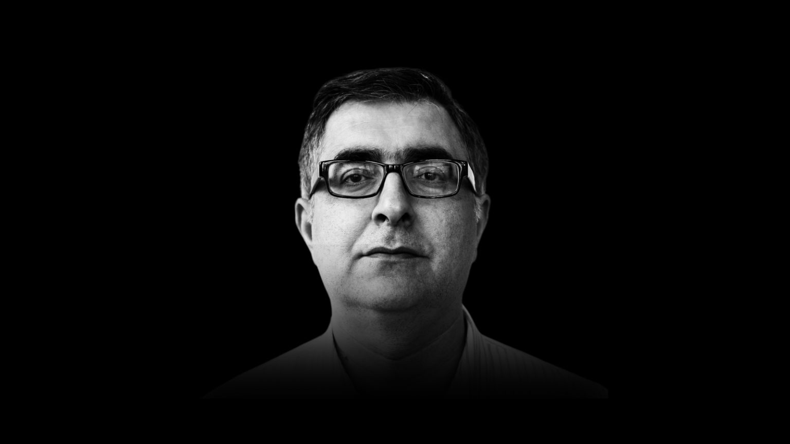 Black and white portrait of a man in glasses looking forward against a dark background.