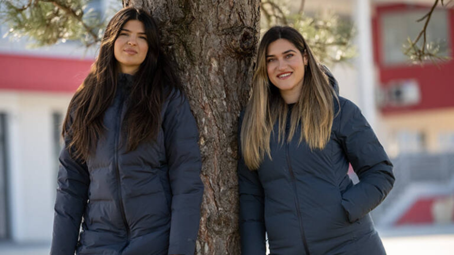 Two young women in dark puffer jackets lean against a tree trunk, smiling.