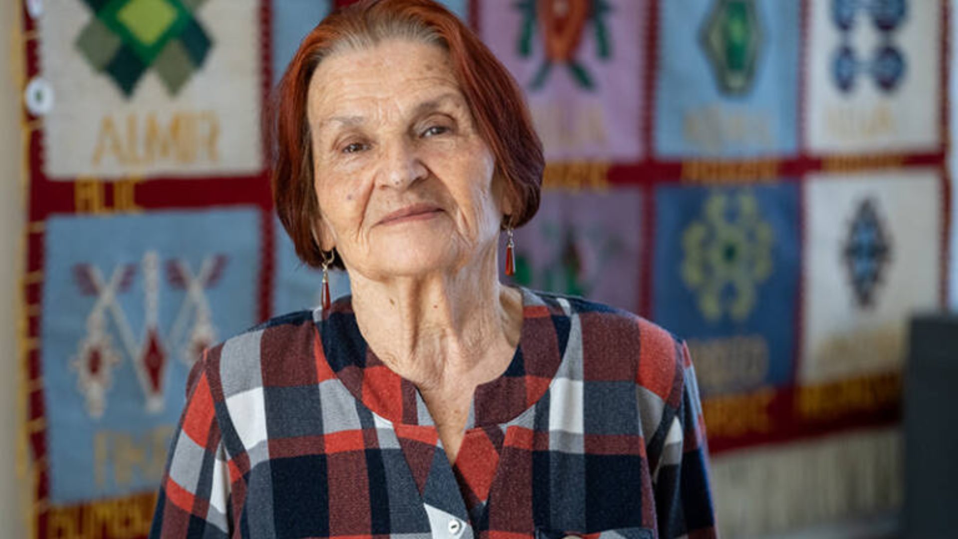 Close-up of an elderly woman with red hair and a plaid shirt, smiling subtly in front of a patterned tapestry.