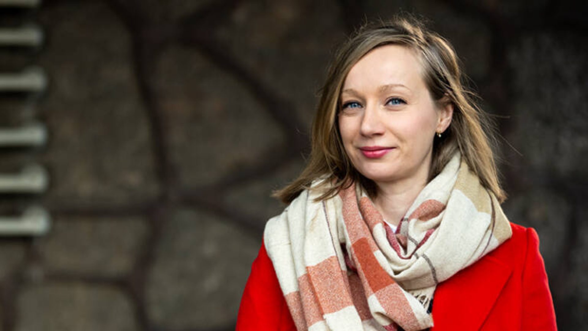A smiling woman with blonde hair and blue eyes, wearing a red coat and a plaid scarf, looking at the camera.
