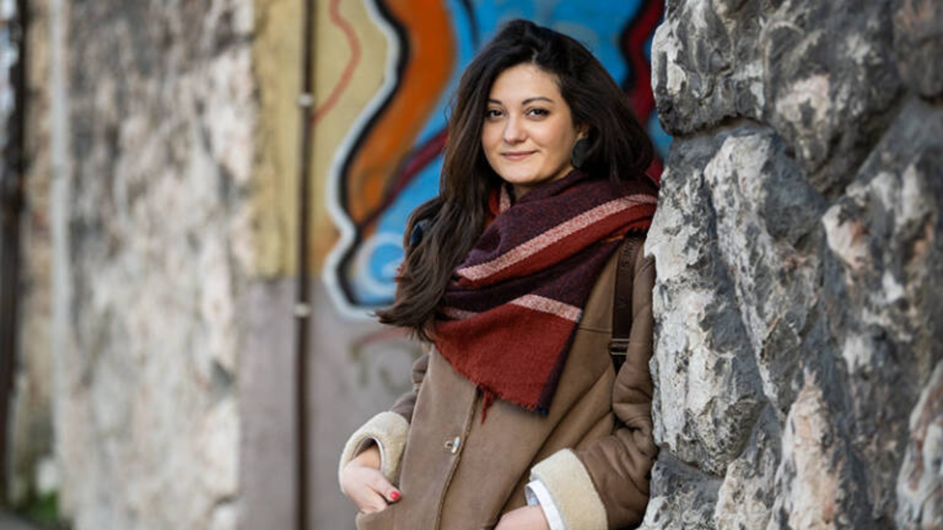 A young woman with long dark hair, wearing a brown coat and scarf, smiles while leaning against a stone wall.