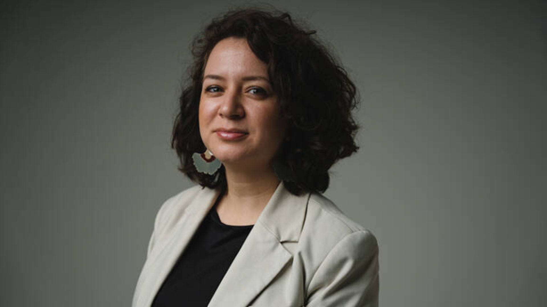 A woman with dark, curly hair and light-colored blazer smiles faintly against a plain grey background.