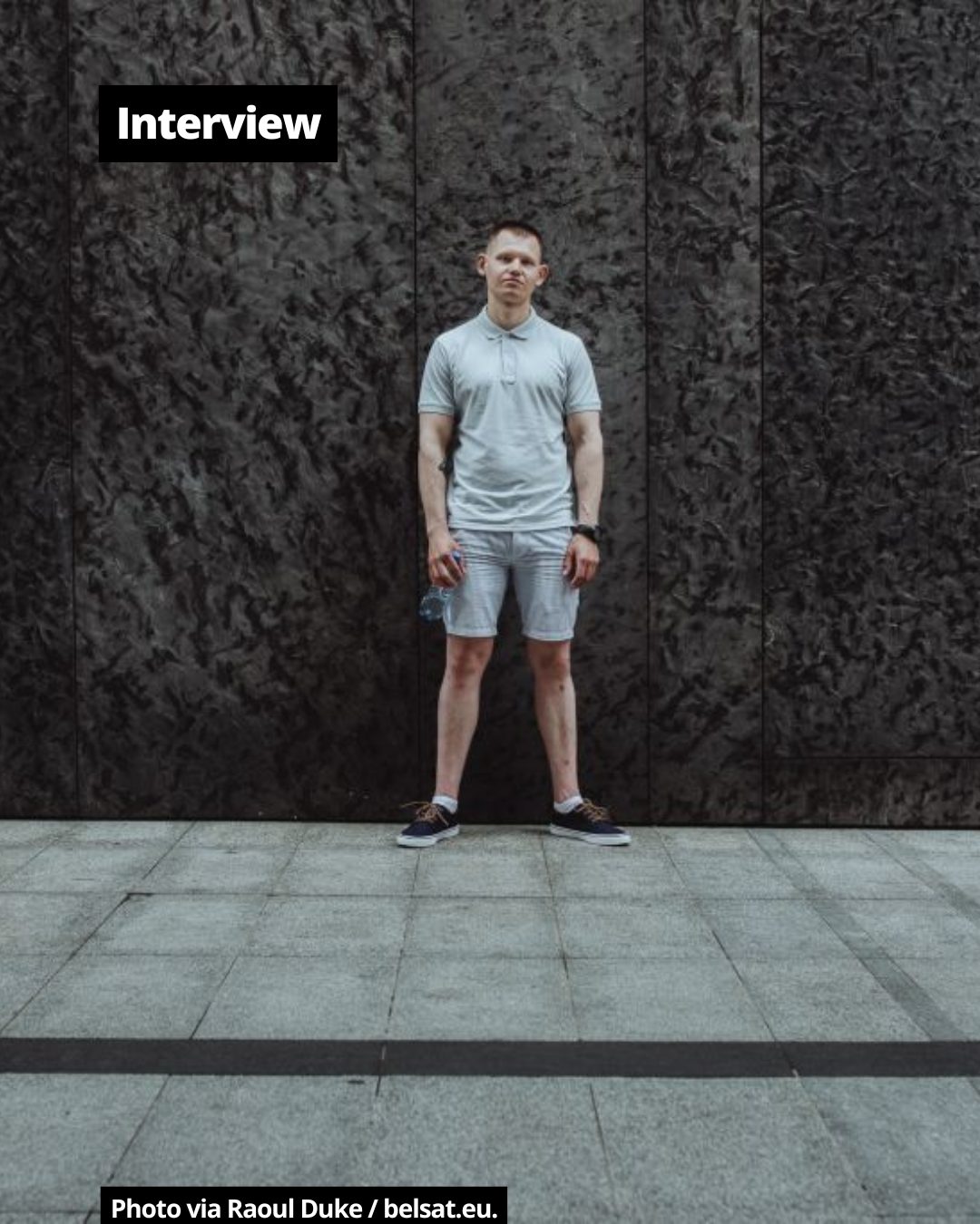 A young man stands facing forward, wearing a light polo shirt and shorts against a dark, textured wall.