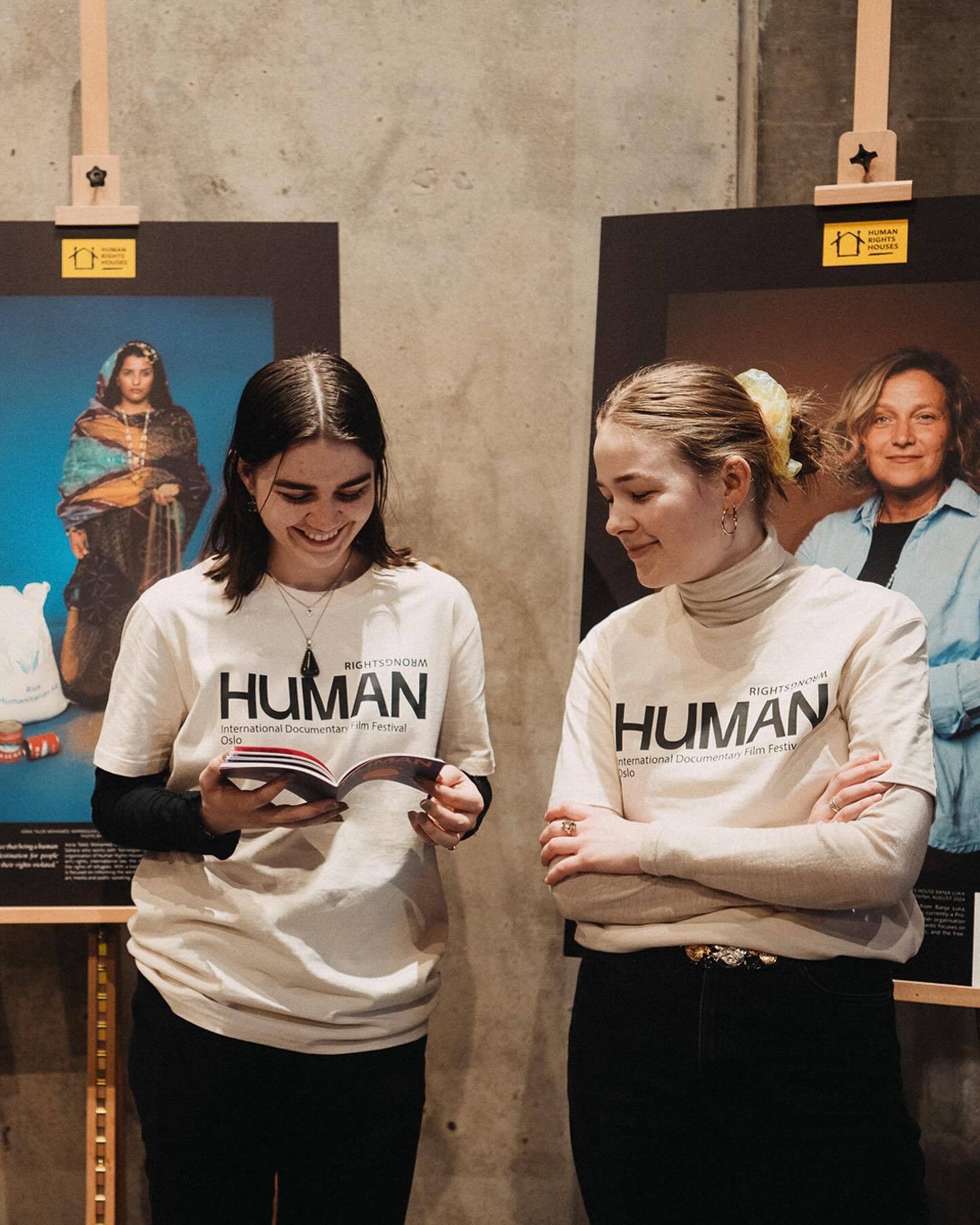 Two smiling women in "HUMAN" T-shirts at a film festival, one holding a book, next to exhibition posters.