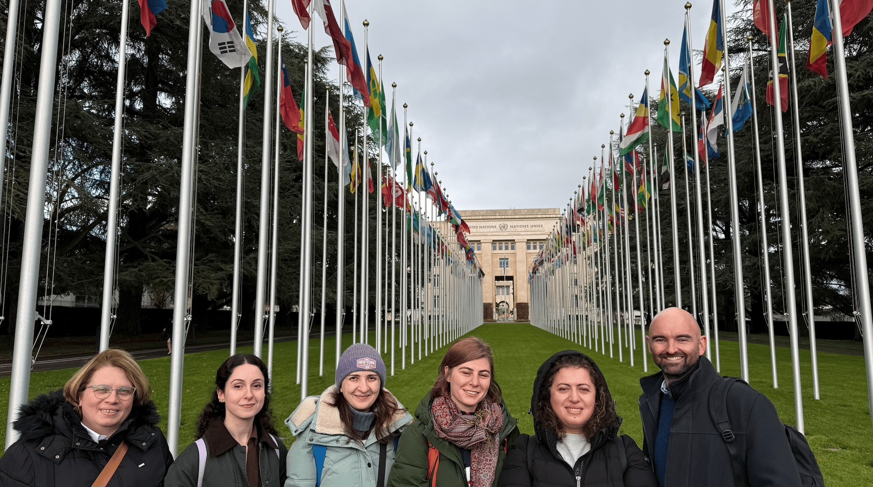 Six people smiling in front of the United Nations building, flanked by rows of international flags.