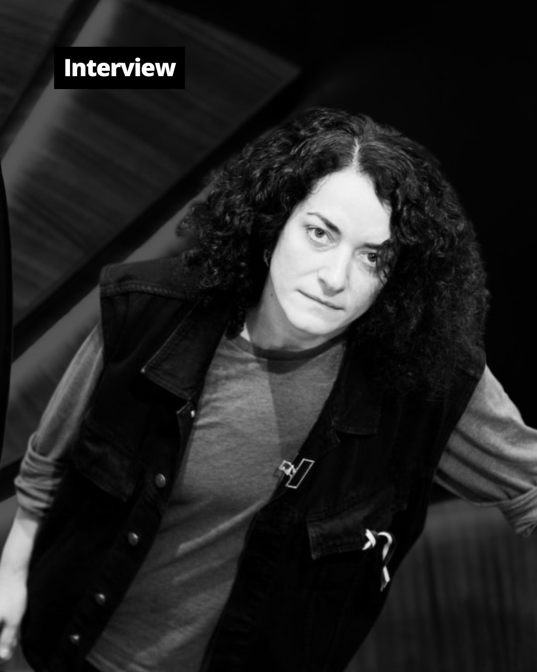 Monochrome portrait of a person with curly hair and a vest, looking directly at the camera.