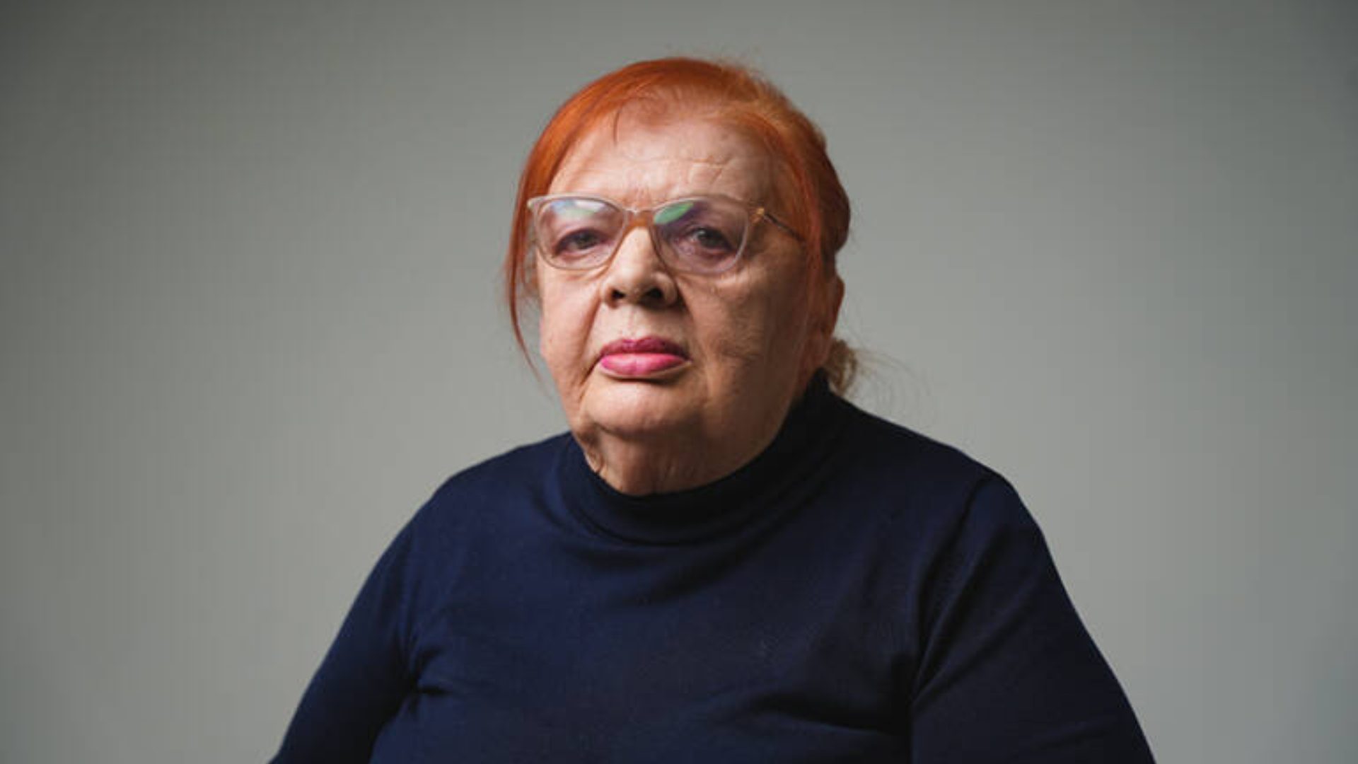 An older woman with red hair and glasses wearing a dark blue turtleneck, looking directly at the camera.