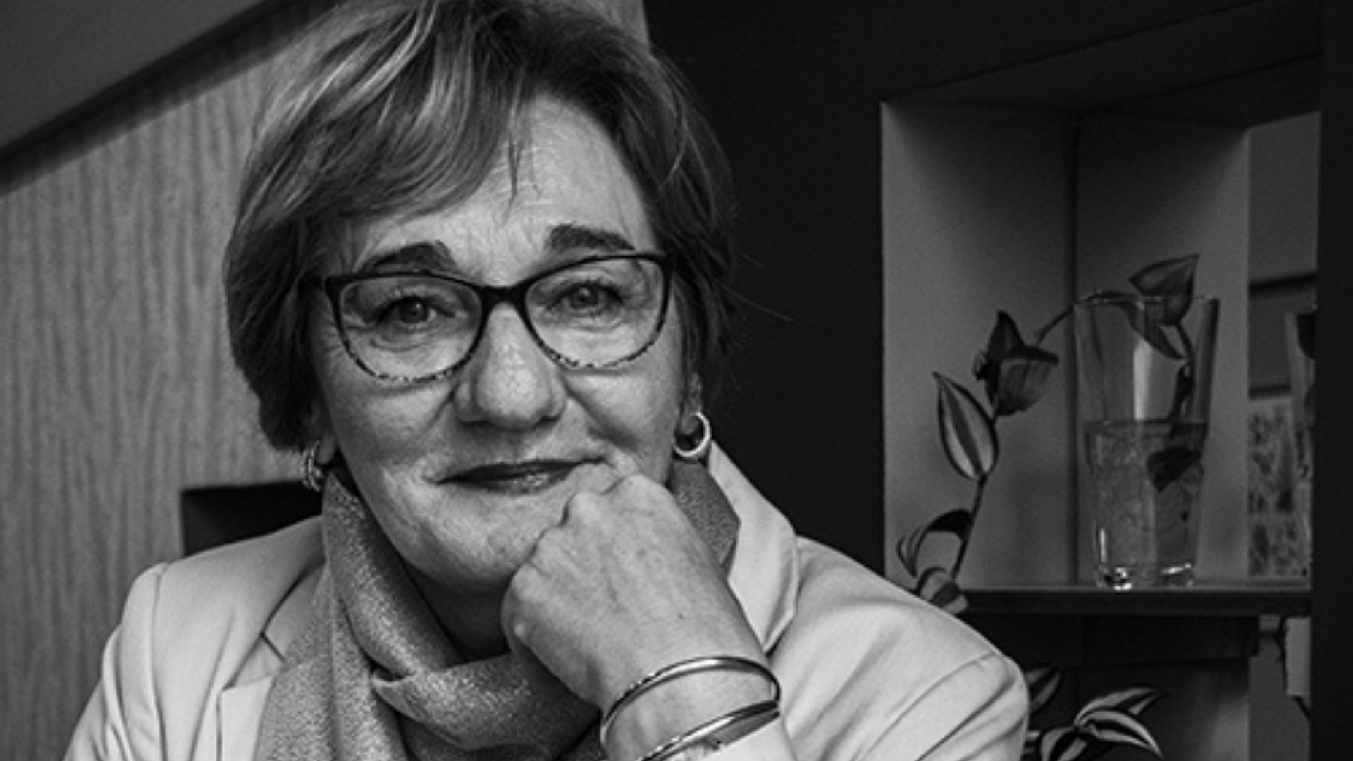 Black and white portrait of a woman with glasses, smiling subtly, with her hand on her chin.