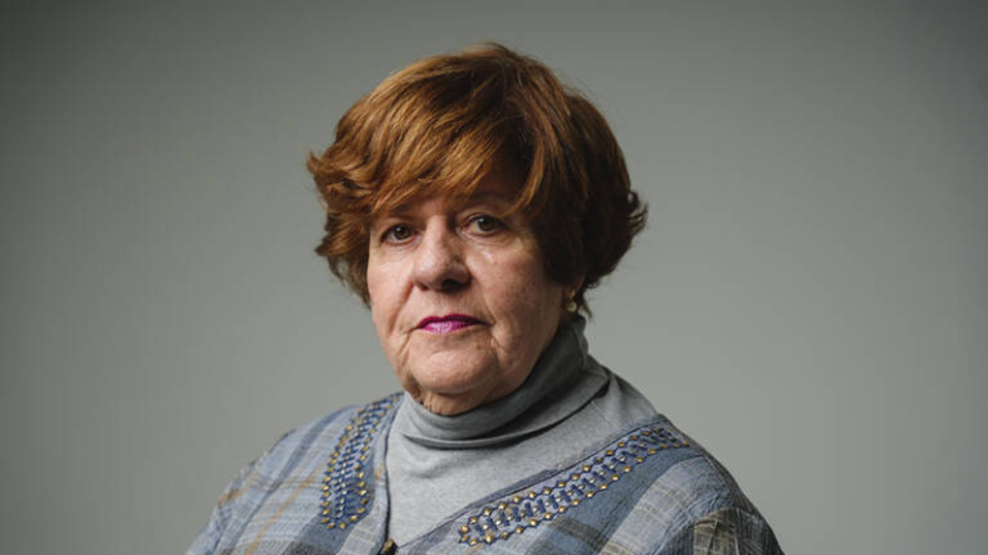 Headshot of an older woman with short red hair and a neutral expression, wearing a gray turtleneck.