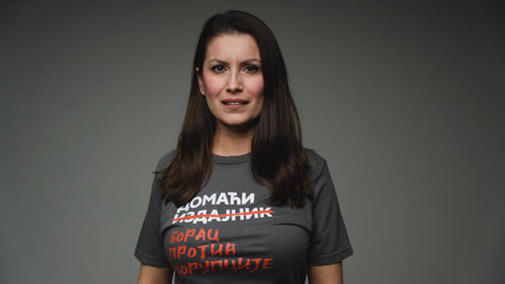 A woman with long dark hair and a serious expression wears a grey t-shirt with red Cyrillic text.