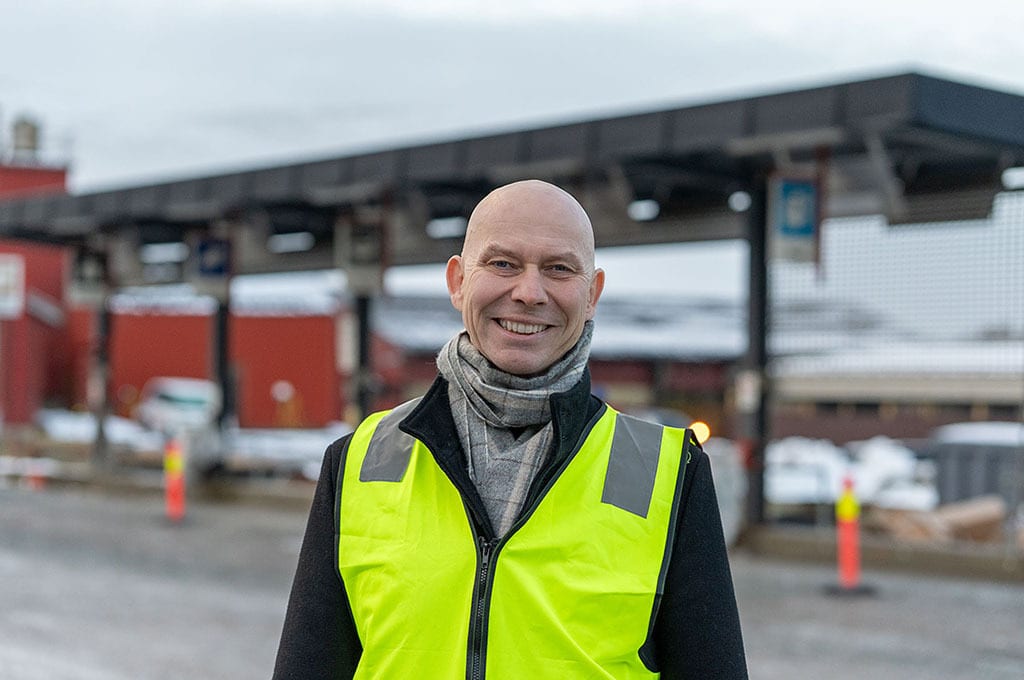 Smiling bald man in yellow high-vis vest and scarf, outdoors at an industrial site.