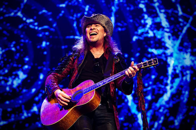 Smiling musician in cowboy hat & red jacket plays guitar.