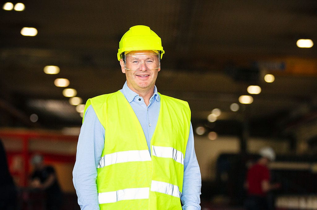 Smiling man in a yellow hard hat, safety glasses, and high-visibility vest.