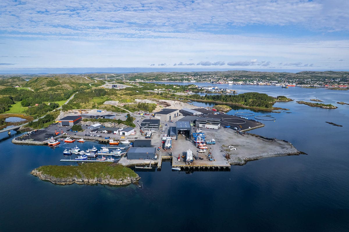 Aerial view of a coastal industrial port with numerous boats, buildings, aquaculture pens, and hilly terrain.