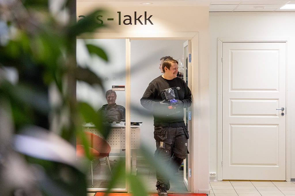 Man in work clothes steps from a doorway smiling, another man smiles from behind glass. Foreground foliage.