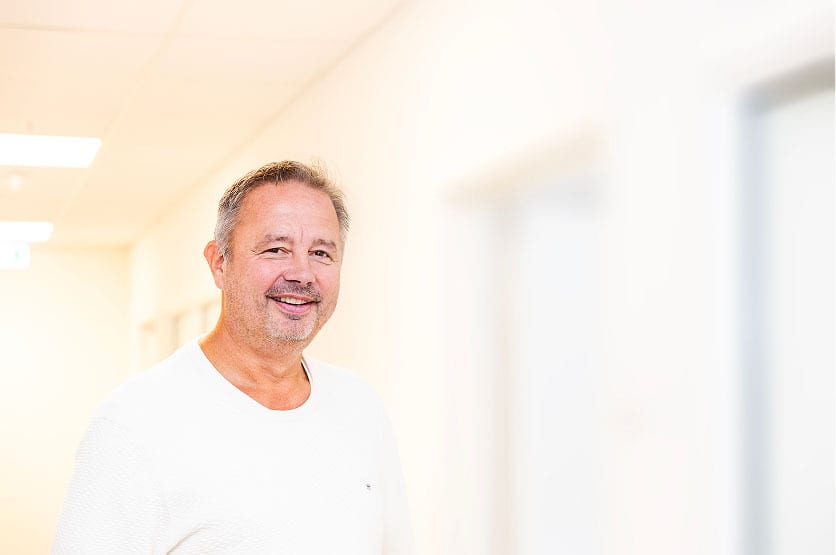 Smiling man with graying hair wearing a white t-shirt in a bright hallway.