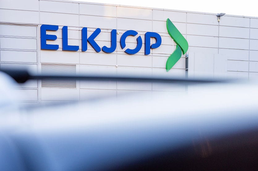 Blue "ELKJOP" sign and green logo on a white building, with blurry foreground.