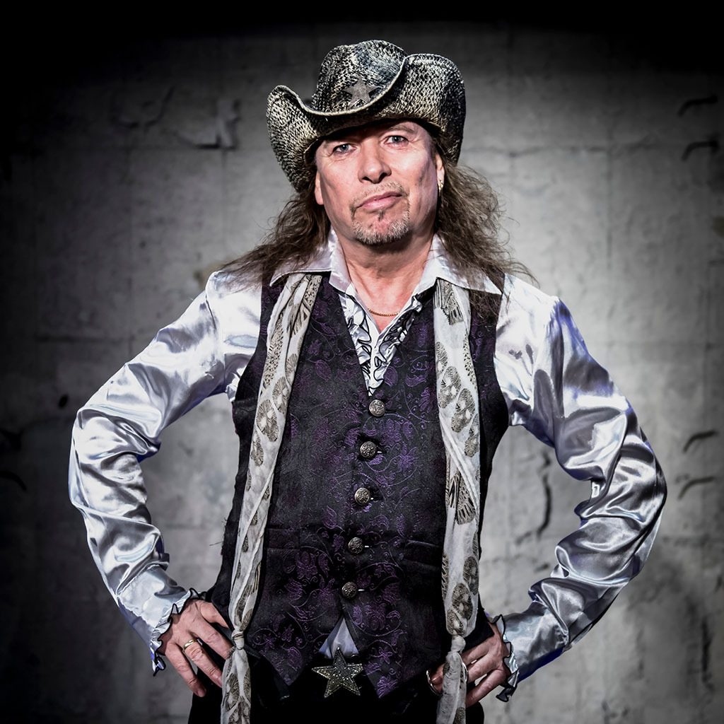 Man in a cowboy hat, long hair, satin shirt, vest, scarf, hands on hips, looking at camera.